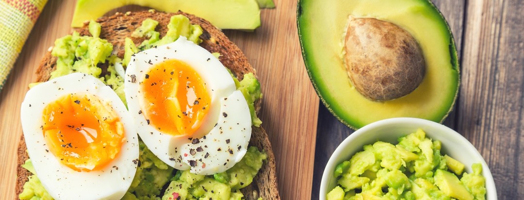 A soft boiled egg is on top of mashed avocado on a piece of toast with half an avocado and more mashed avocado to the right of it.