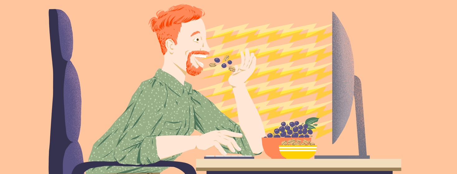 An energetic-looking man sits forward in an office chair facing a computer screen. One hand types on a keyboard, one tosses grapes and walnuts into his mouth.