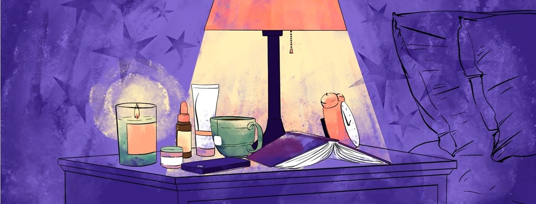 A lamplit nightstand has a collection of items on top of it - skin care products, a lit candle, a mug of tea, a cellphone, a book, and an alarm clock.
