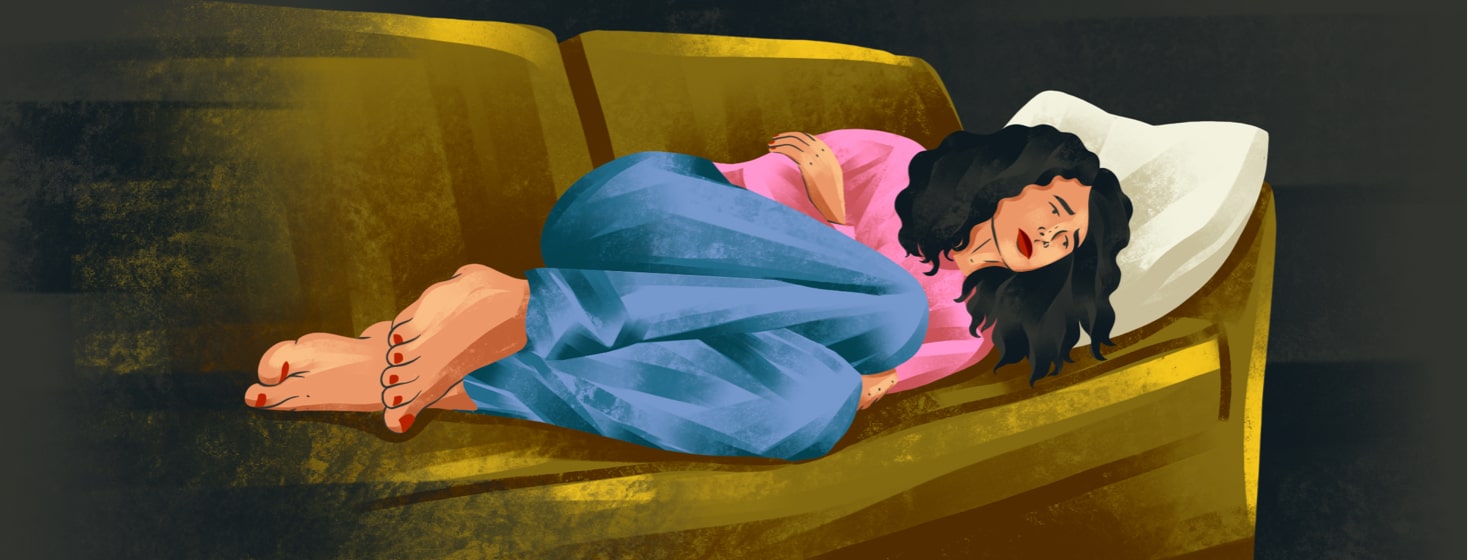 alt=a woman lies on her side on a couch, clutching her stomach in discomfort